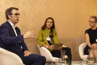 Drei Personen sitzen auf beigen Stühlen auf einem Podium und tauschen sich aus. Der Mann in dunklem Anzug spricht gerade und macht eine Handbewegung. Die beiden Frauen hören ihm zu. Den Hintergrund ziert eine beige-gemusterte Wand.