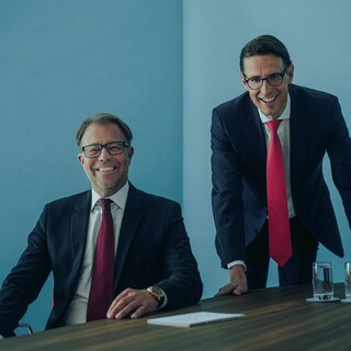 Zwei lachende Herren in Anzug an einem Tisch.