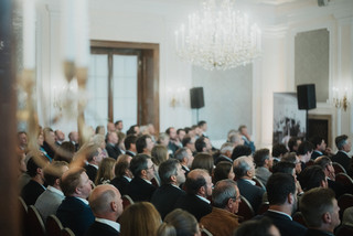 Elegant gekleidetes Publikum sitzt in einem historischen Saal unter einem Kronleuchter.