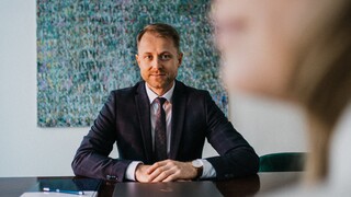 Am Tisch sitzt ein Mann im blauen Anzug, die Hände auf dem Tisch.