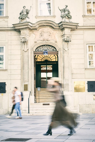 Äußere Fassade eines historischen Gebäudes, einer Niederlassung des Bankhaus Spängler. Im Vodergrund ist ein Paar ersichtlich, ebenfalls eine Frau, die eilig geht.