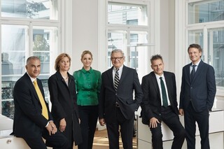 6 Personen (2 Frauen, 4 Männer) in einem hellen Büro. 5 Personen tragen dunkle Kleidung, 1 Frau trägt ein dunkelgrünes Oberteil.