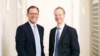 Zwei Männer stehen in einem hellen Büro. Beide tragen einen schwarzen Anzug mit weißem Hemd. Ein Mann hat dunkle Haare und trägt eine hellblaue Krawatte, der andere hat helle Haare und trägt eine dunkelblaue Krawatte. Beide tragen eine Brille und lächeln freundlich.